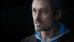 Retrato de Oleksii Sivak, quien lleva barba corta y bigote, mira hacia la izquierda de la cámara y viste una sudadera con capucha azul claro debajo de una chaqueta negra.