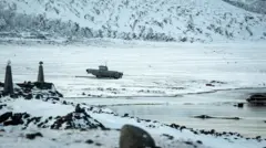 Frozen water and a snow-covered landscape in Greenland, with a military-looking vehicle pictured in the middle distance