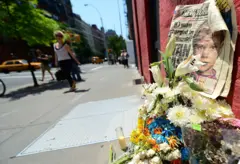 Un arrume de flores sobre el cual se apoya un periódico en el que se ve una foto de Etan