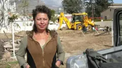 Uludağ, bir arazi aracının yanında duruyor, arkasında bir iş makinesi var, kameraya bakıyor, elleri ceplerinde. Tarihi bilinmiyor. 