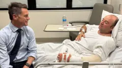 A picture inside a hospital room. On a grey hospital bed with white sheets lies Mr Ahmed, a balding man wearing a white t-shirt with his left arm and wrist in a cast. Chris Minns sits at the end of the bed smiling at him, wearing a light blue shirt, dark blue tie and black trousers.