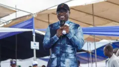 Romuald Wadagni wear blue dress, black face cap and hold microphone