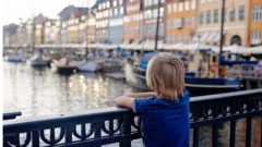 طفل لون شعره أصفر، يقف أمام نهر ويشاهد المراكب المصطفة