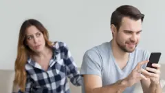 muškarac gleda u telefon, iza njega je žena koja ga gleda ljuto i razočarano