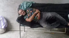 Lázaro Romero León mira a cámara tumbado en una silla alargada, con jeans gris oscuros y una camiseta estampada, en un patio de cemento en Tapachula, Chiapas, México.