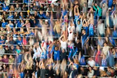 Gradería en un estadio llena de personas con algunos terminando de levantarse, otros levantados con los brazos en alto, y al lado, otros preparándose para hacerlo.