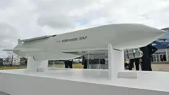 Bir stand üzerinde duran uzun beyaz bir füze ve çevresinde insanlar görülüyor. Üzerinde “storm shadow/scalp” yazıyor; arka kısmında kanatlar ve sivri bir burun bulunuyor.