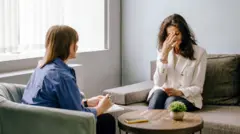 Mujer sentada con la mano en el rostro, afligida por la pena, conversa con una terapeuta que toma nota de la conversación