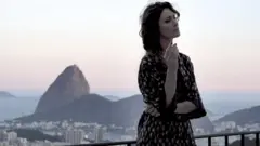 Maria Fernanda Cândido, uma mulher branca de cabelos castanhos cacheados, usa um vestido estampado e posa em uma sacada com o Pão de Açucar ao fundo