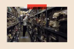 Un hombre con sudadera gris claro saca una pila de archivos de una estantería en una sala de archivos, que está llena de otros archivos