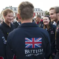 La espalda de la chaqueta del hombre luce una bandera británica y la inscripción "Fuerzas Armadas Británicas". Está de pie frente al príncipe Harry entre una multitud.