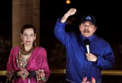 Rosario Murillo con aretes y un collar color marrón, un vestido de flores coloridas y un chal rosado está parada al lado de Daniel Ortega, quien lleva puesta una chaqueta azul y una gorra negra. El mandatario tiene un brazo levantado y con el otro sostiene un micrófono
