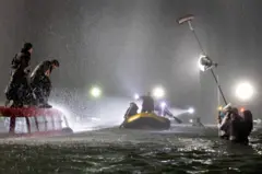 Cena noturna de filmagem na água sob chuva forte: pessoas se equilibram sobre um carro parcialmente submerso enquanto um bote se aproxima, e, ao lado, a equipe opera luz e som dentro da água.