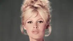 Um retrato de estúdio de Brigitte Bardot usando um vestido azul claro tomara que caia com um penteado loiro estilo colmeia e os lábios ligeiramente entreabertos