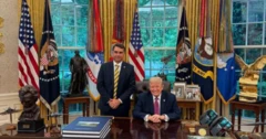 Foto mostrando Flávio Bolsonaro, em pé, ao lado de Donald Trump, sentado, no salão oval da Casa Branca, repleto de bandeiras, porta-retratos e uma janela ao fundo