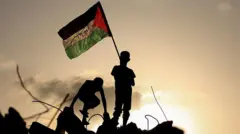 Crianças palestinas brincam enquanto uma delas acena com uma bandeira nacional palestina em pé sobre os escombros de um prédio destruído no campo de Bureij, em Gaza. Elas estão em silhueta contra o pôr do sol que ilumina o fundo da imagem