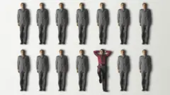 Una persona de camisa bordó y pose relajada entre filas de hombres de traje gris y pose rígida