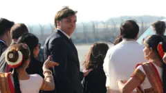 Em área aberta, Bolsonaro olha para trás em direção à câmera, enquanto é acompanhado por assessores e mulheres com trajes típicos na recepção ao presidente brasileiro