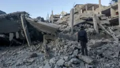 Вайрон бўлган Нусайрат қочқинлар жамлоғи бинолари олдида турган эркак