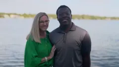Stacey y Marven frente a la cámara frente a un lago. Ambos sonríen. Marven lleva una camiseta gris con cuello. Stacey tiene el pelo largo y rubio, gafas y una blusa verde brillante fruncida.