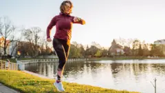 A woman takes a run next to a pond in a UK town. She is looking at her watch and there is early morning light on her face