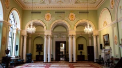The court room in the Bank of England which has decorated green walls, an elaborate gold, green, white and gold ceiling and a patterned rug with white columns and arches on the far side of the room