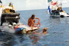 Dos automóviles de los años 70 flotan en el mar. Sobre sus techos hay banderas, velas y botes auxiliares. Un hombre rubio (Marco Amoretti) está en el agua con la mano apoyada en el capó del coche. Otro hombre de cabello castaño y barba (Marco De Candia) está en cuclillas sobre el capó del mismo vehículo mientras manipulan una cuerda atada a él.