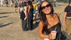 Lauren Macpherson está em um festival. Ao fundo, é possível ver uma multidão de pessoas. Ela segura uma bebida rosa em um copo plástico com a mão esquerda e tem uma bolsa preta no ombro. Usa óculos de sol pretos, tem cabelos castanhos e longos e sorri para a câmera