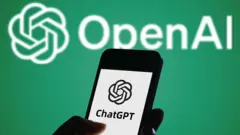 Uma mão segurando um telefone com o logotipo do ChatGPT na tela. Ao fundo, vê-se uma tela verde com o logotipo da OpenAI.