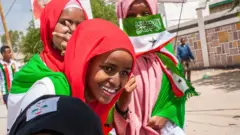 Trois jeunes femmes souriantes portant des foulards rouges et roses et tenant un drapeau du Somaliland et d'autres insignes - Hargeisa, 2018