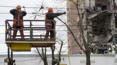 Двоє енергетиків стоять на висоті, ремонтують електричні дроти біля зруйнованого будинку. Позаду них розтрощена будівля. 