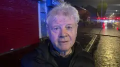 Um homem parado na estrada à noite. Ele tem cabelos brancos e está olhando diretamente para a câmera.