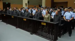 Pessoas em fila em um tribunal, cercadas por agentes de segurança.