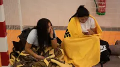 Dos mujeres dentro de una portería de fútbol sala, en manga corta pero abrigafas en las piernas con mantas. Alrededor tienen algunos enseres en mochilas. 
