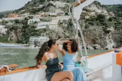 Duas mulheres, visivelmente relaxadas e felizes, desfrutam de um passeio de barco no Mar Mediterrâneo. Ao fundo, avista-se a famosa cidade de Positano, na Itália.