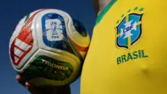 Pessoa com a camisa oficial da seleção brasileira segurando a bola oficial da Copa do Mundo de 2026