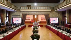 Sağda ve solda uzun kırmızı örtülü masalarda oturan komisyon üyeleri karşılıklı görünüyor. Ortada çiçekler sırayla meclis başkanı Numan Kurtulmuş'un kürsüsüne uzanıyor. Kürsünün arka planında büyük tablolar ve 6 adet Türk bayrağı var. 