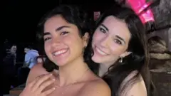 Duas garotas sorrindo para a câmera. À esquerda, a garota tem cabelo escuro na altura dos ombros. Ela está usando uma blusa jeans azul e um colar prateado. A garota à direita tem cabelo escuro e longo. Ela está usando uma blusa jeans azul e brincos prateados. A foto foi tiradabetpix365 bonus 20um bar com longas mesasbetpix365 bonus 20madeira ao fundo.