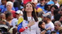 María Corina Machado con una mano en el pecho, mirando hacia arriba, vistiendo una camiseta con la bandera venezolana, rodeada de simpatizantes durante un acto político.