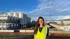 Letícia Lemos Martins veste um colete amarelo refletivo e sorri, com a fábrica da Heineken em Passos, Minas Gerais, ao fundo. Ela tem cabelos ondulados compridos e veste uma camiseta preta por debaixo do colete