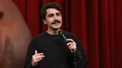 Deniz Göktaş'ın Harbiye'de sahnelediği gösteri sırasında kırmızı bir perde önünde konuşuyor. Bıyıklı ama genç bir görüntüsü var. Bir elinde mikrofon tutuyor.