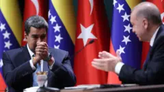 Maduro solda, Cumhurbaşkanı Erdoğan sağda duruyor. Maduro ellerini birbirine kavuşturmuş, Erdoğan alkışlıyor. İki liderin arkasında Türk ve Venezuela bayrakları var.