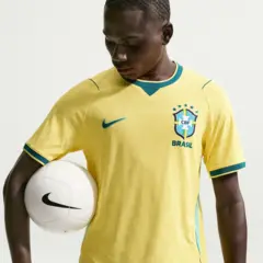 A imagem mostra um jovem segurando uma bola de futebol branca da Nike debaixo do braço enquanto veste a camisa amarela da Seleção Brasileira. O uniforme tem detalhes em verde e azul, com o escudo da CBF e cinco estrelas no peito, além do logotipo da Nike do outro lado. O jogador olha para baixo, em direção à camisa.