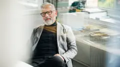 Homem, possivelmente baby boomer, sentado no escritório com óculos pretos, blusa preta e jaqueta cinza, com as mãos sobre as pernas