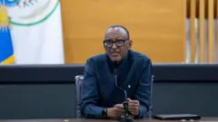 Perezida w'u Rwanda Paul Kagame arimo kureba imbere muri 'camera', yicaye mu ntebe y'umusego w'umukara. Yambaye indorerwamo n'ishati yijimye y'amaboko maremare, afashe ikaramu mu biganza byombi. Imbere ye ku meza hari indangururamajwi ntoya iriho agapamba k'umukara. Ifoto yo ku wa kane, ku itariki ya 27 Ugushyingo (11) mu 2025, ubwo yari ari mu kiganiro n'abanyamakuru, mu biro bye byitwa Village Urugwiro, i Kigali, mu Rwanda.
