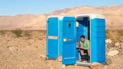Homem sentado no vaso sanitário de um banheiro móvel no deserto. A porta está aberta e ele mexe no celular