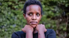 Agather Atuhaire ahagaze yambaye umupira w'amaboko magufi w'umukara urimo utudomo tujya kuba umweru, agaragaza inkovu ku maboko yombi iruhande rw'aho ibiganza bitangirira, nyuma yo kurekurwa n'abategetsi ba Tanzania. Ifoto yo ku itariki ya 23 Gicurasi (5) mu mwaka wa 2025, ari hanze y'ibitaro by'i Kampala, muri Uganda. 