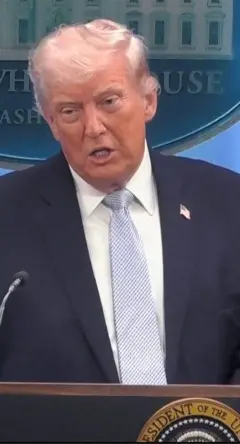 ලා නිල් පාටින් කොටු වැටුණු සුදු පැහැති ටයි එකක් පැළඳ, සුදු පැහැති කමිසයක් ඇඳ කළු කෝට් එකකින් සැරසී සිටින ඩොනල්ඩ් ට්‍රම්ප් මෙහි සිටී.