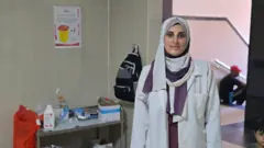 القابلة ياسمين أحمد ترتدي معطفها الأبيض، وبجوارها طاولة، عليها معدات طبية