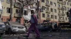 Девушка в розовом идет на фоне поврежденного здания.